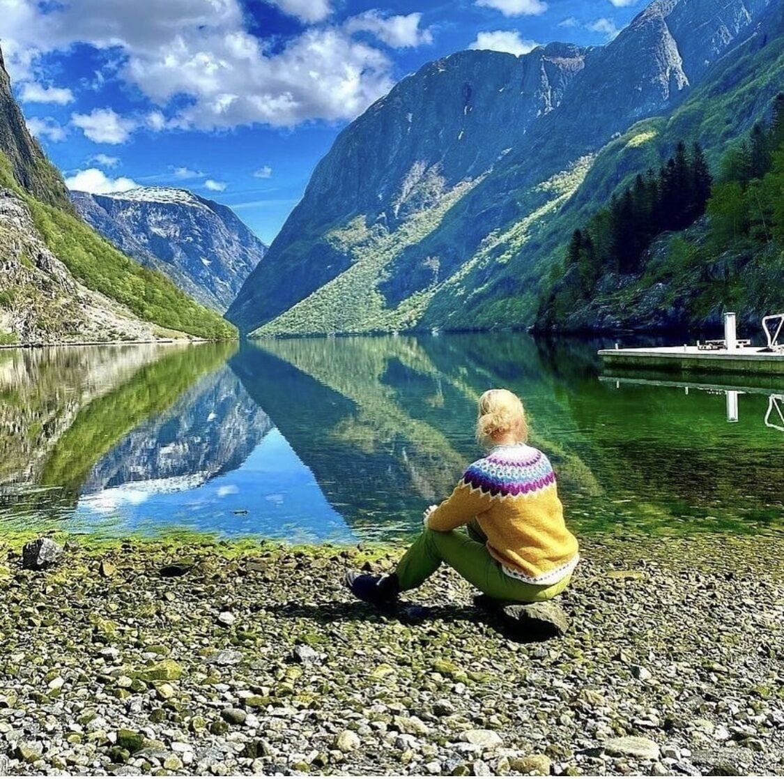 Une personne assise au bord d’un lac paisible, avec des montagnes en arrière-plan.