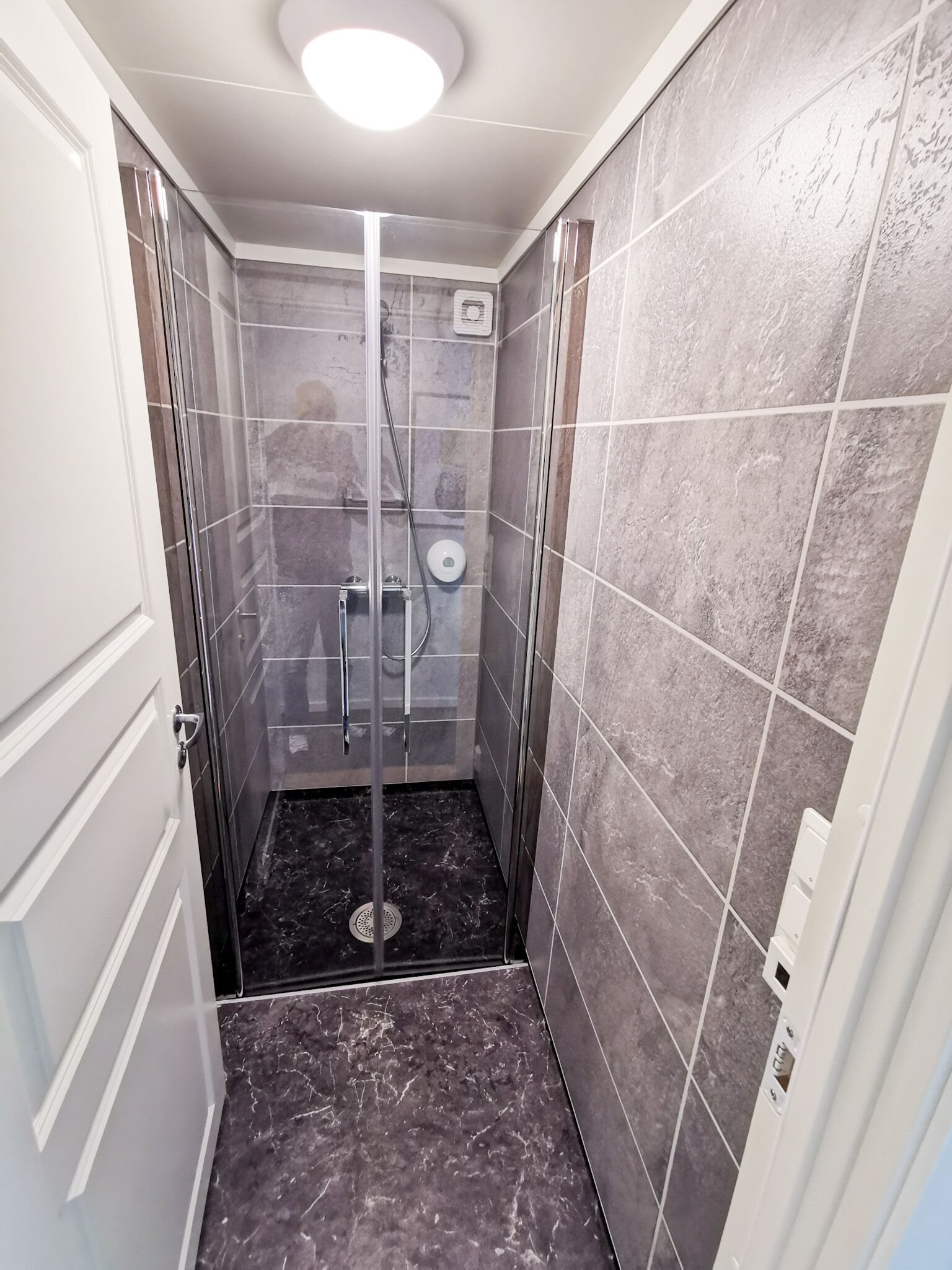 A small, enclosed shower stall with a glass door, a handheld showerhead, and a wall-mounted soap dispenser.