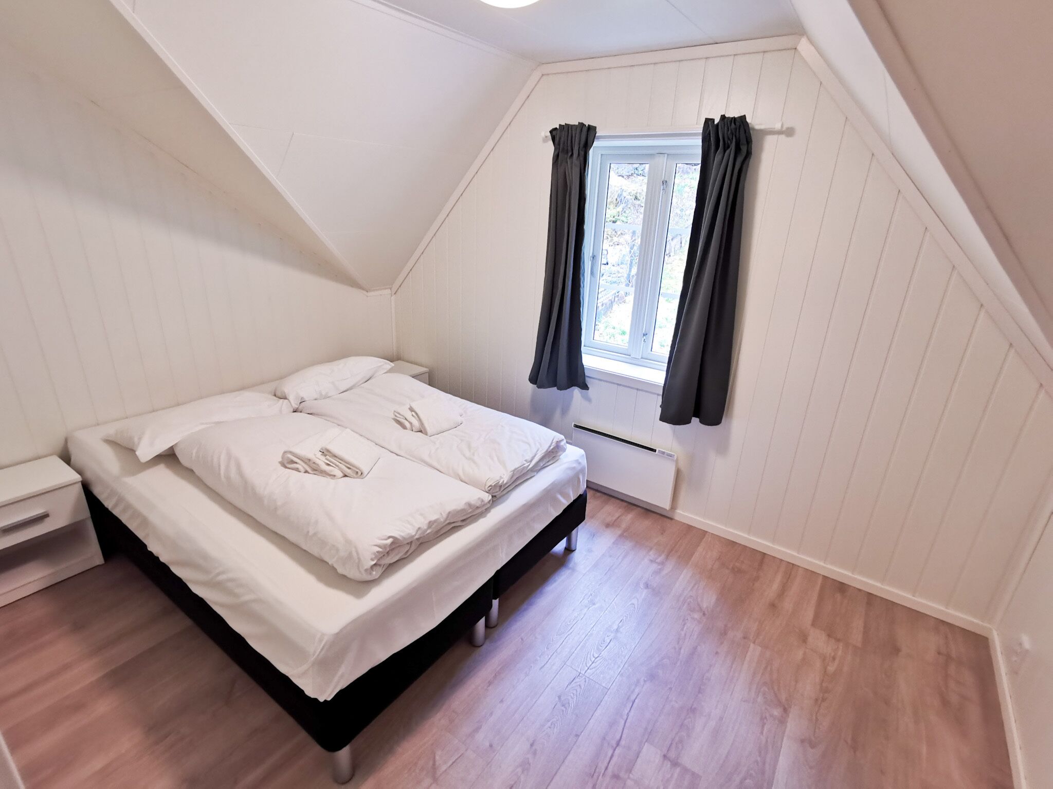 A bedroom with a bed, a nightstand, a window with curtains, and a radiator.