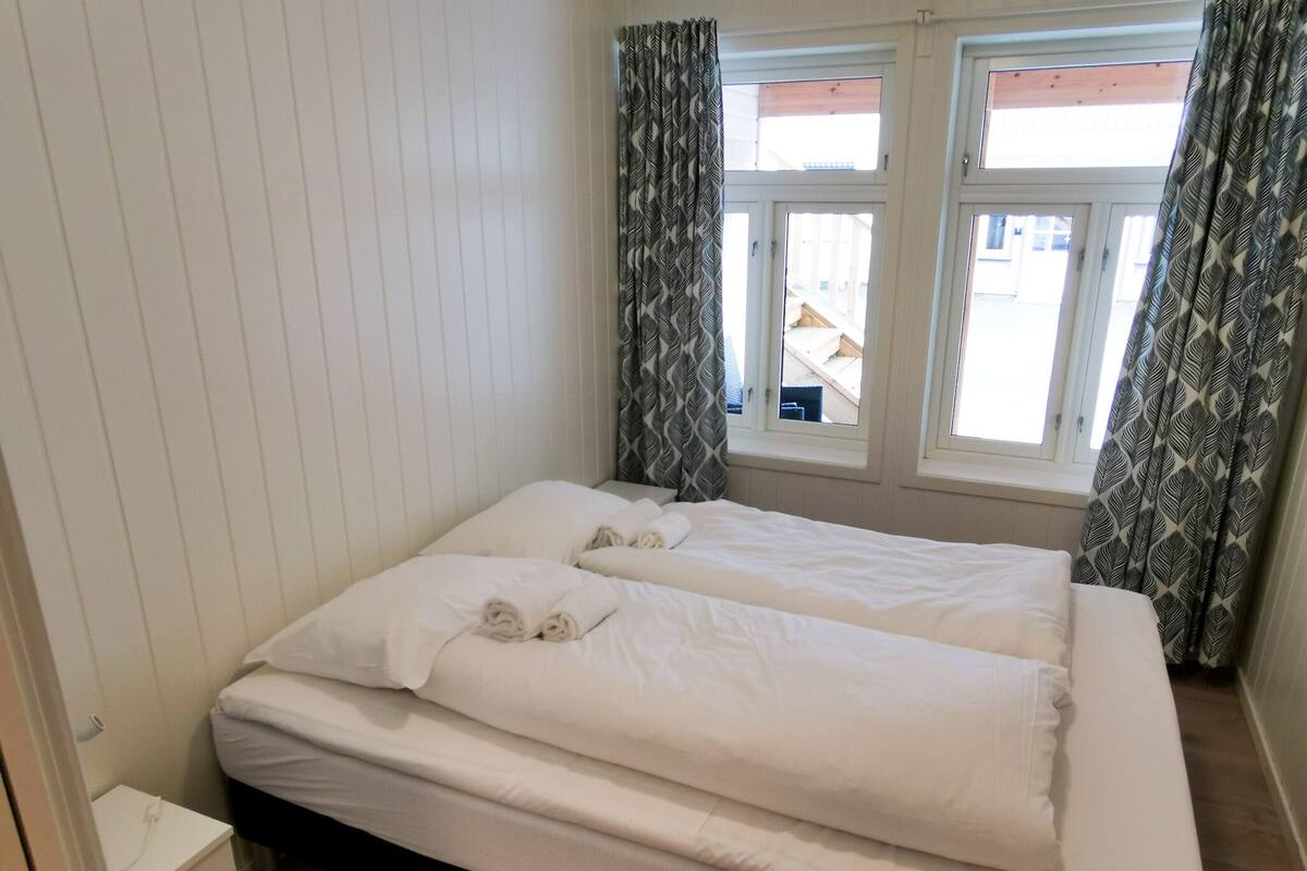 Una cama doble con ropa de cama blanca, una mesita de noche y una ventana con cortinas estampadas.