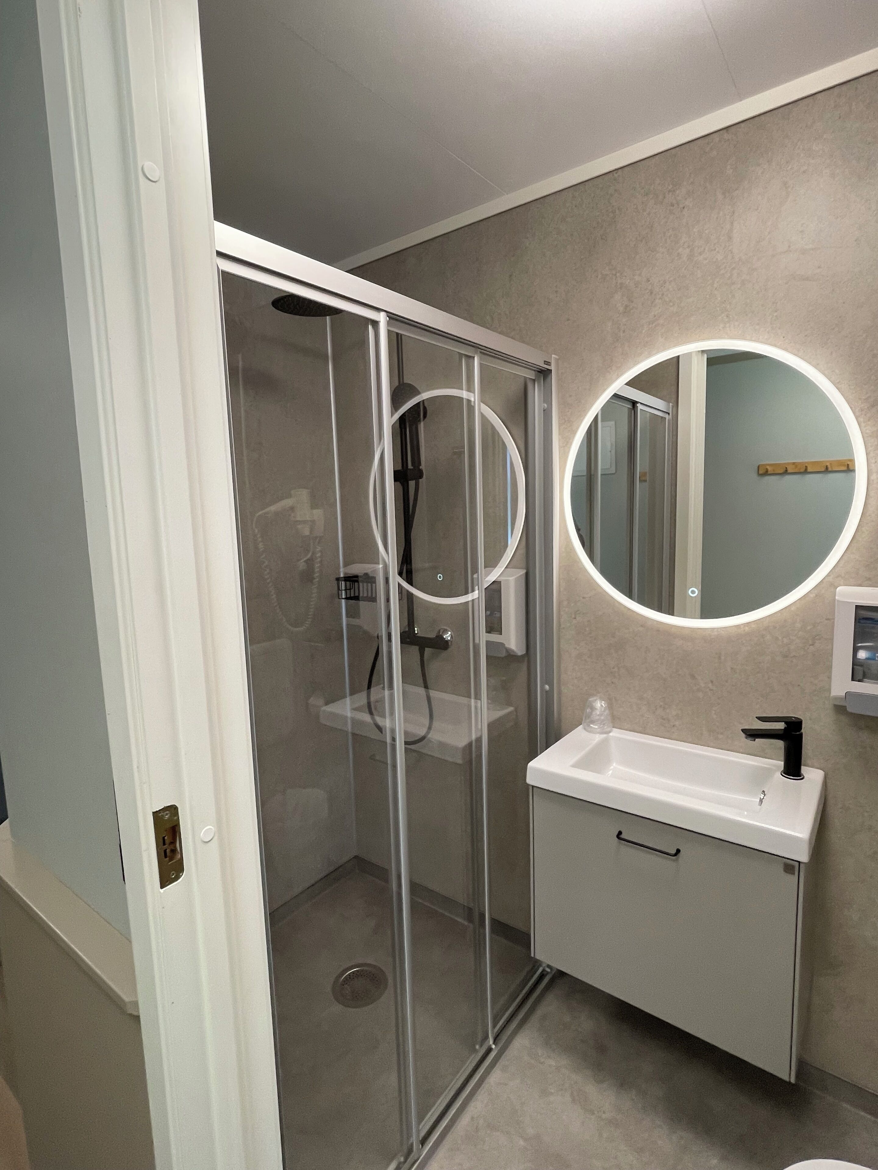 Ein modernes Badezimmer mit einer gläsernen Dusche, einem weißen Waschbecken mit schwarzem Wasserhahn und einem runden Spiegel mit Beleuchtung.