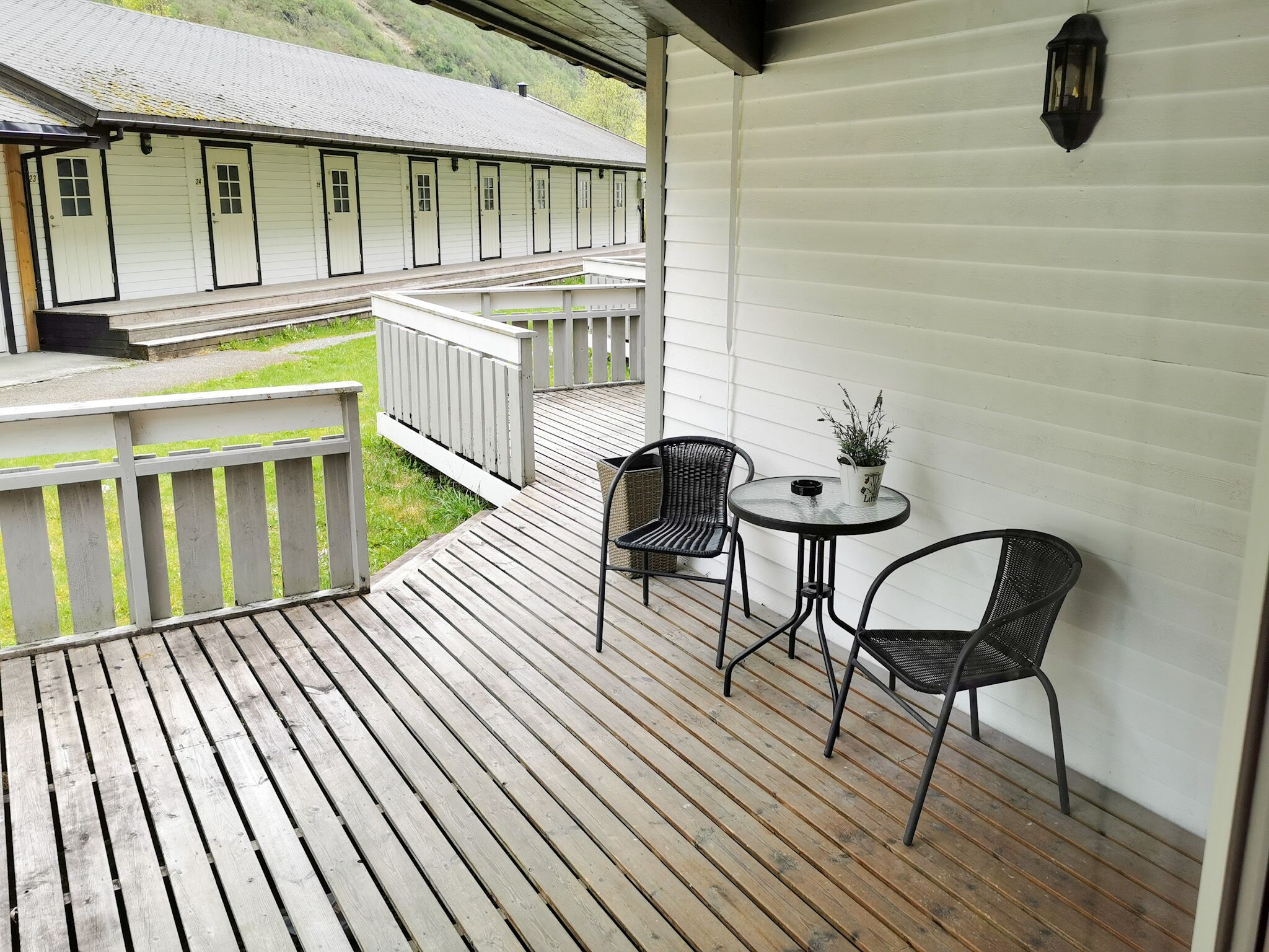 A wooden deck with two wicker chairs and a small table, overlooking a row of white buildings with numbered doors.