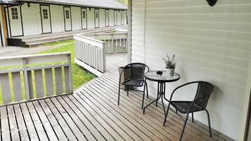 A wooden deck with two wicker chairs and a small table, overlooking a row of white buildings with numbered doors.