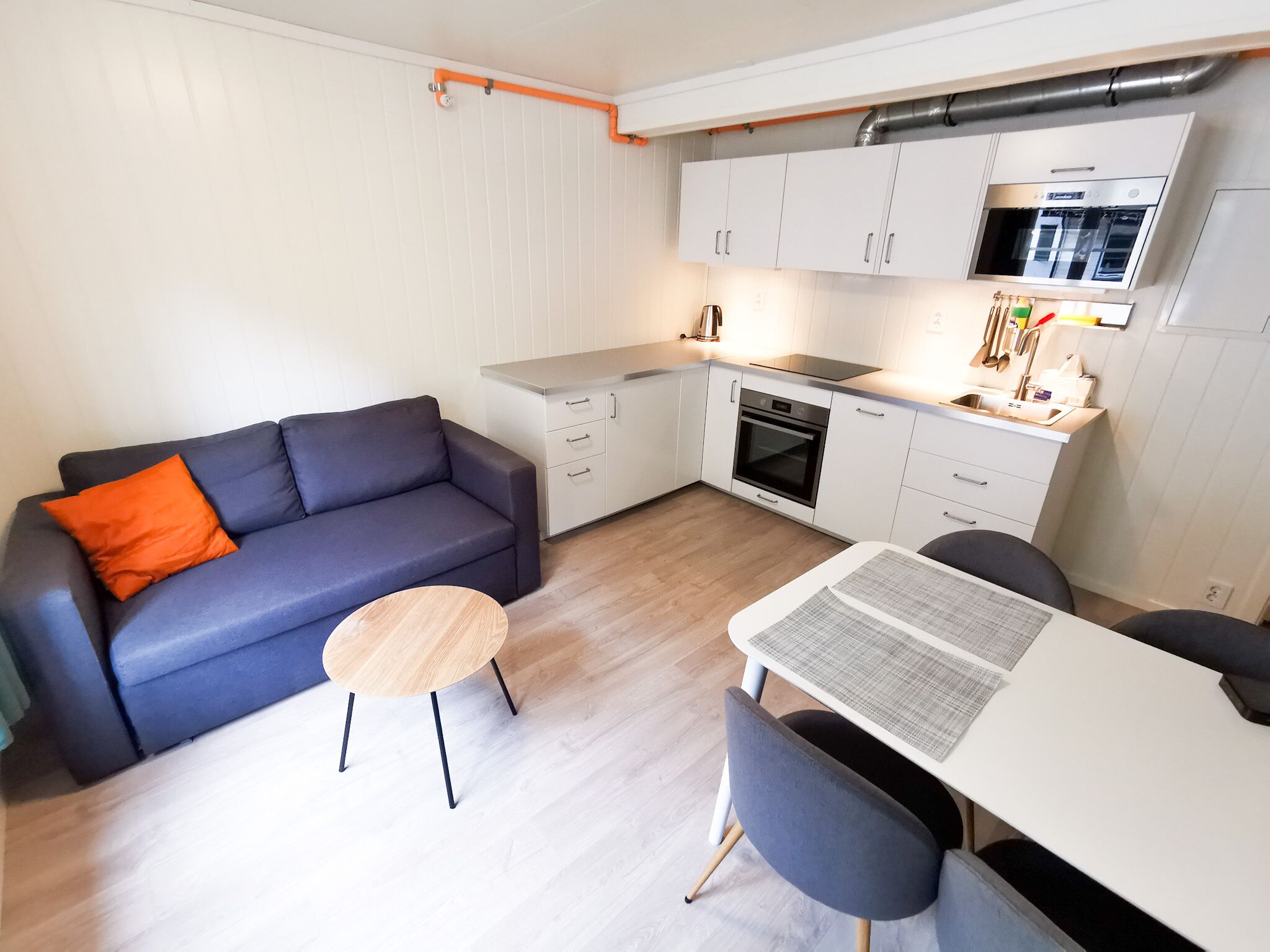 A compact living space with a sofa, a small round table, a dining table with chairs, a kitchenette with a microwave, and a stove.
