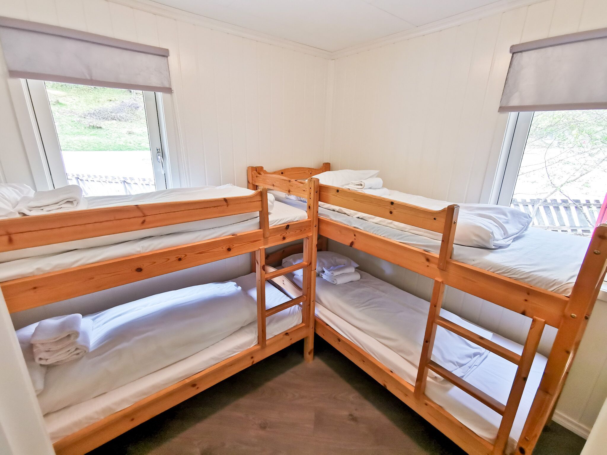 Een kamer met hoekligging, drie stapelbedden, wit beddengoed en ramen met jaloezieën.