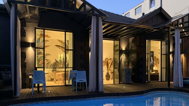 A modern outdoor pool area with a wooden deck, glass doors leading to a lounge area with chairs and a table, and a clear view of the pool. - Mountview Guest House (Cape Town)