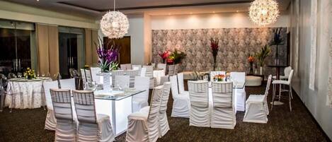 A spacious dining area with round tables and chairs, elegant chandeliers, and a patterned wall in the background.