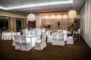 Uma ampla área de jantar com mesas e cadeiras redondas, elegantes lustres e um papel de parede estampado ao fundo.