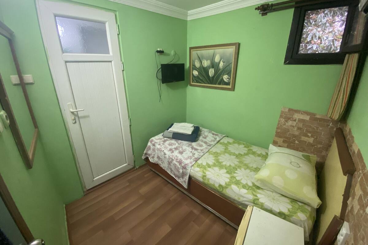 Ein kleiner Raum mit grünen Wänden, einem Einzelbett, einer weißen Tür und einem kleinen Fenster.