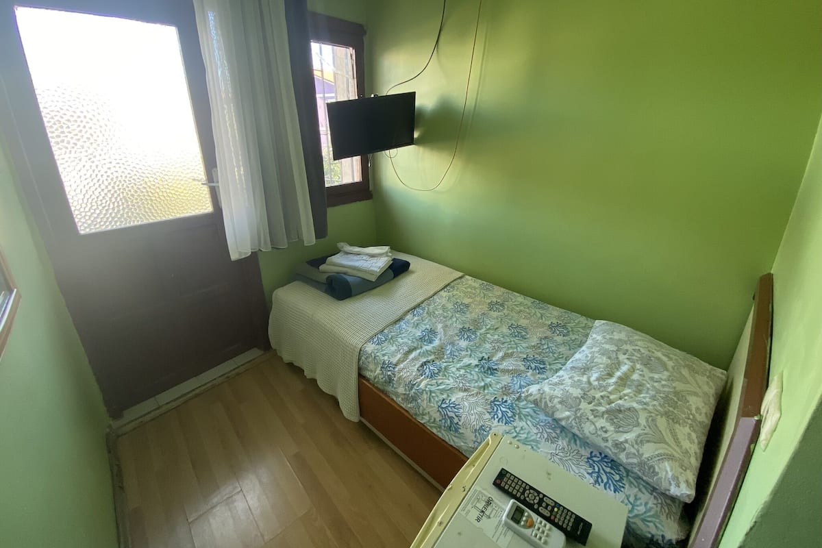 Ein kleines Zimmer mit einem Einzelbett, Holzboden, Bett mit gemustertem Überwurf, Wand-Fernseher und einem Vorhangfenster.