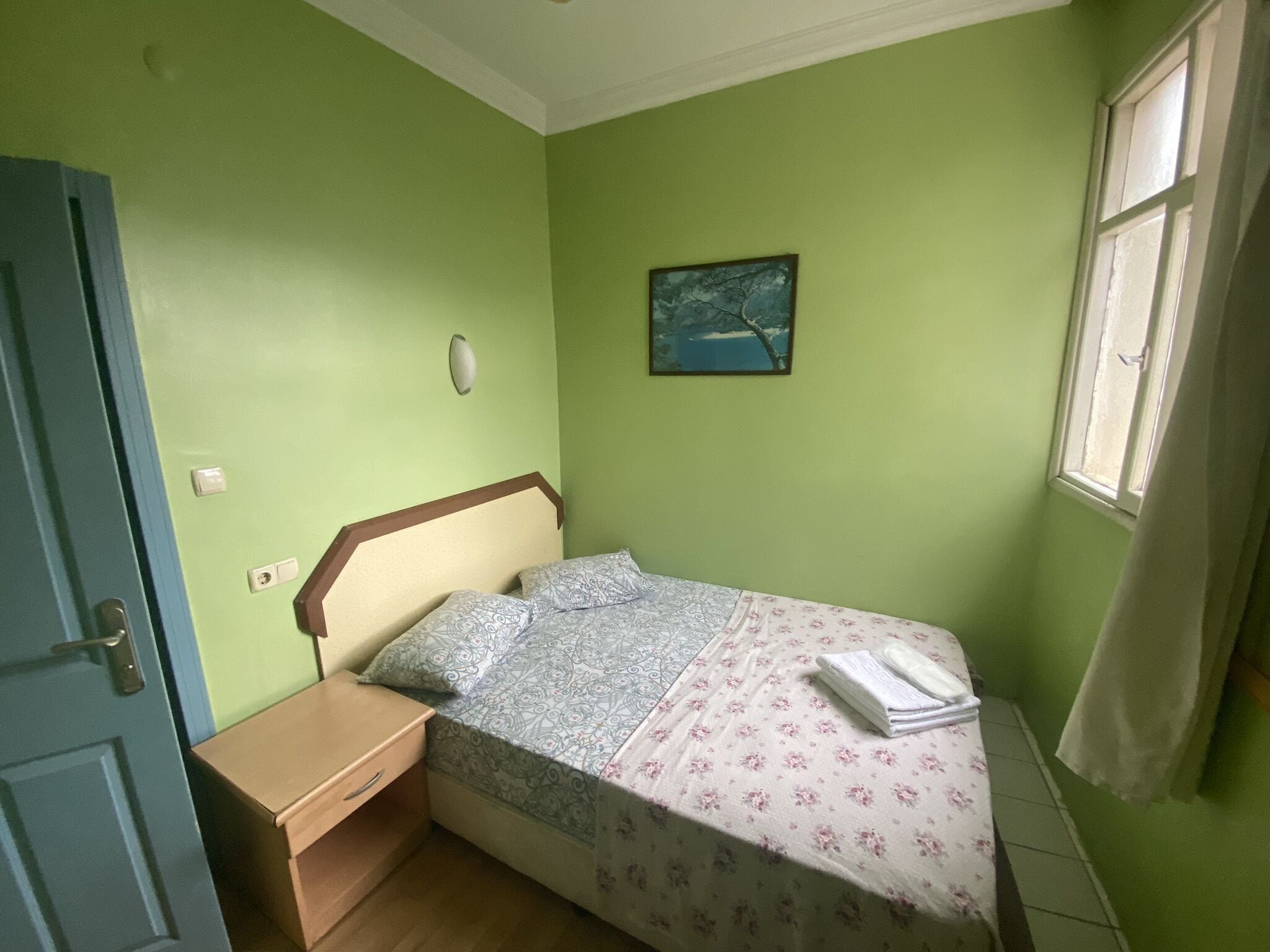 Une petite chambre avec un lit simple, une table de chevet en bois, un lit recouvert d’une literie à motifs, un tableau accroché au mur et une fenêtre avec des rideaux.