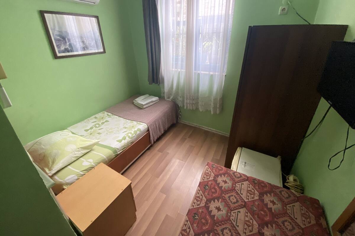 Ein kleines Zimmer mit einem Einzelbett, einem Holzschrank, einer weißen Klimaanlage, einem gerahmten Bild an der Wand und einem Fenster mit durchsichtigen Vorhängen.
