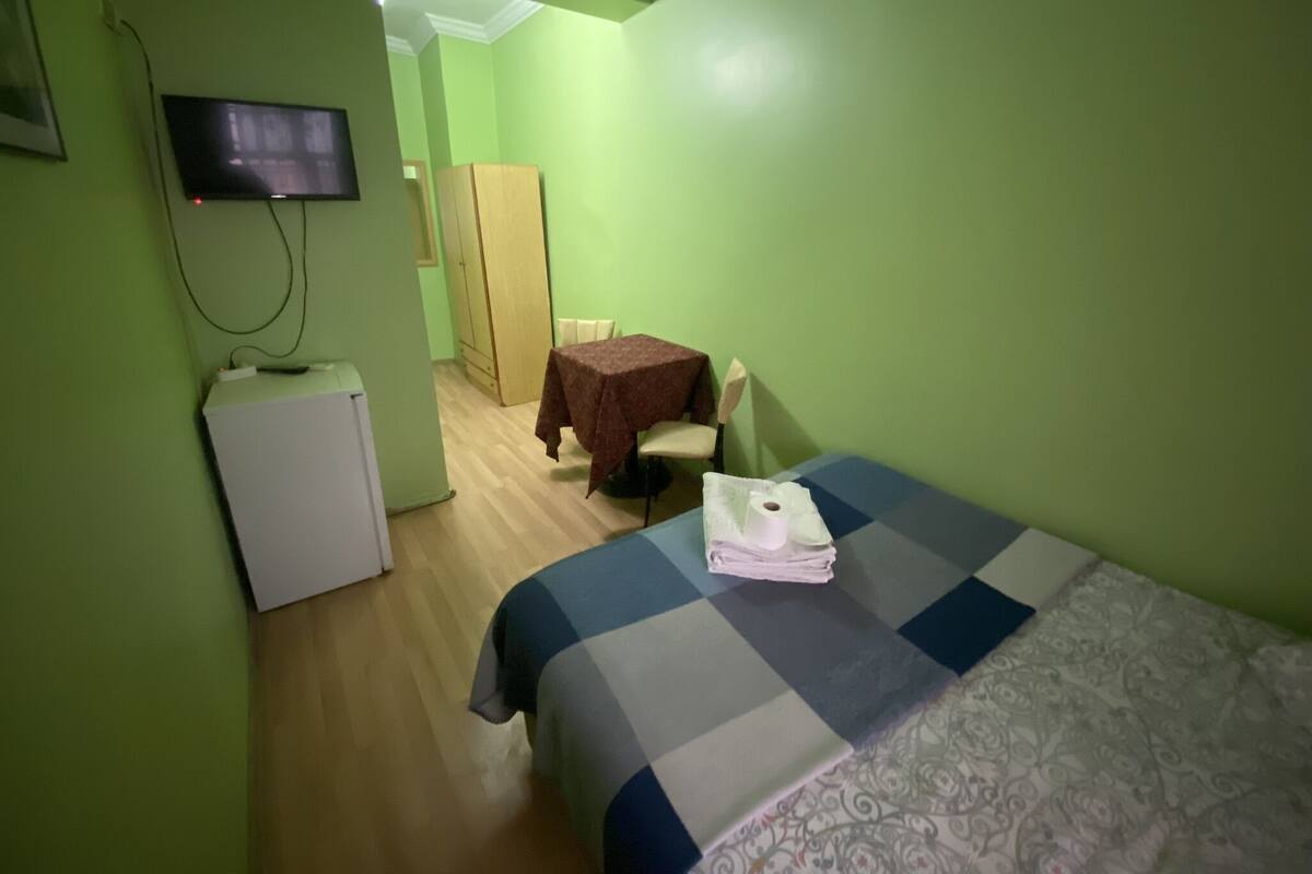 Ein kleiner Raum mit grünen Wänden, ausgestattet mit einem Bett, einem Tisch mit Tischdecke, einem Stuhl, einem an der Wand befestigten Fernseher und einem kleinen Kühlschrank.
