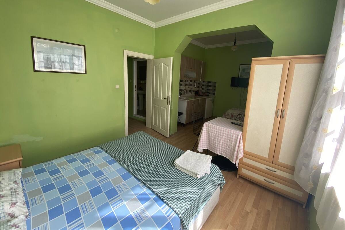 Ein Schlafzimmer mit Bett, kleinem Tisch, Kleiderschrank und einer Küchenzeile im Hintergrund.