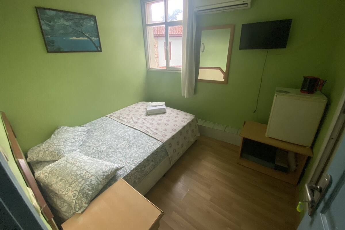 Ein kleines Zimmer mit einem Einzelbett, Holzboden, einem Bett mit gemusterten Betttextilien, einem kleinen Tisch, einem an der Wand befestigten Fernseher und einem Fenster mit Blick auf ein Gebäude.