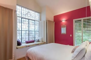 A bedroom with a bed, a window with decorative grills, a red wall, and a mirror.