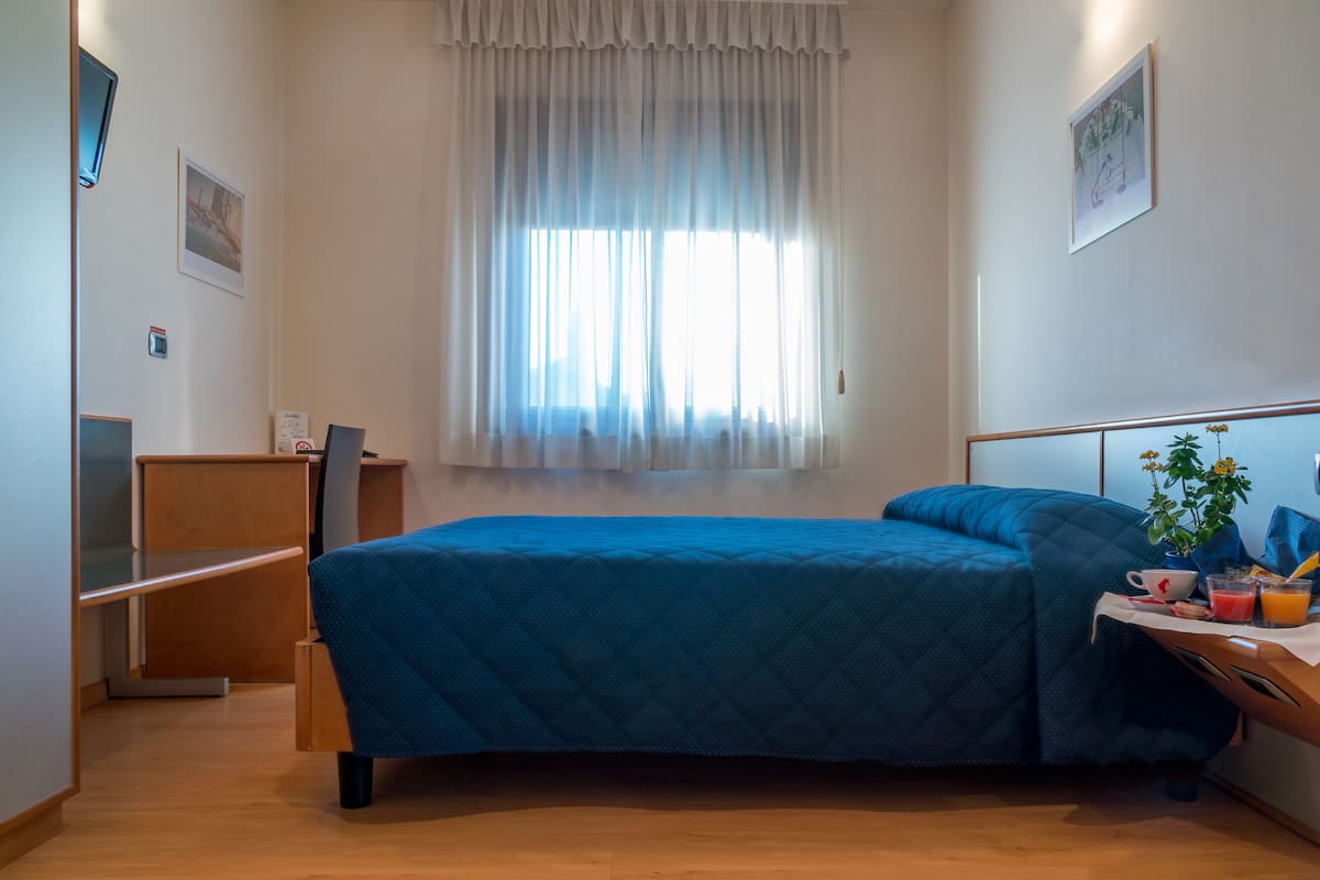 A bedroom with a bed, a desk, a chair, a window with curtains, and a wall-mounted thermostat.