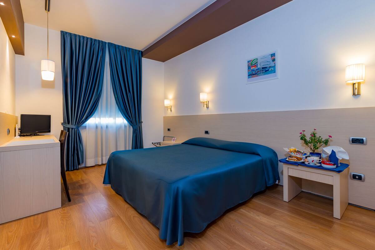 A hotel room with a bed, a desk, a chair, a TV, and a wall-mounted lamp.