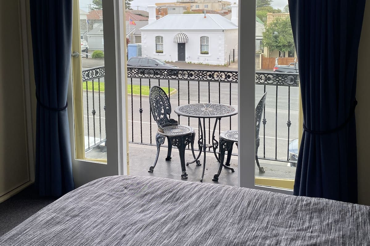 A room with a balcony featuring a table and chairs, a view of the street, and a building in the background.
