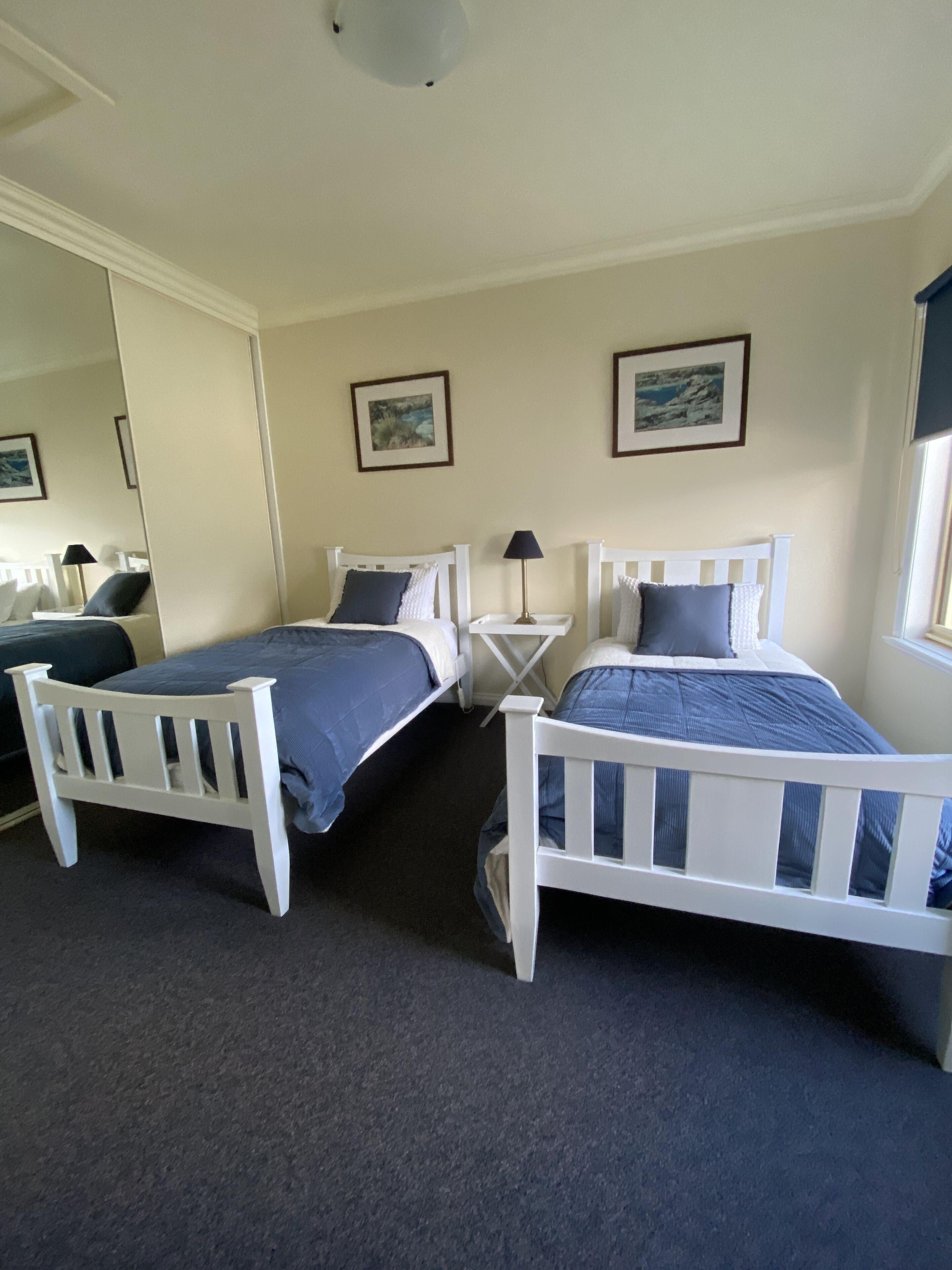 A bedroom with two single beds, a bedside table with a lamp, a large mirror, and framed pictures on the wall.