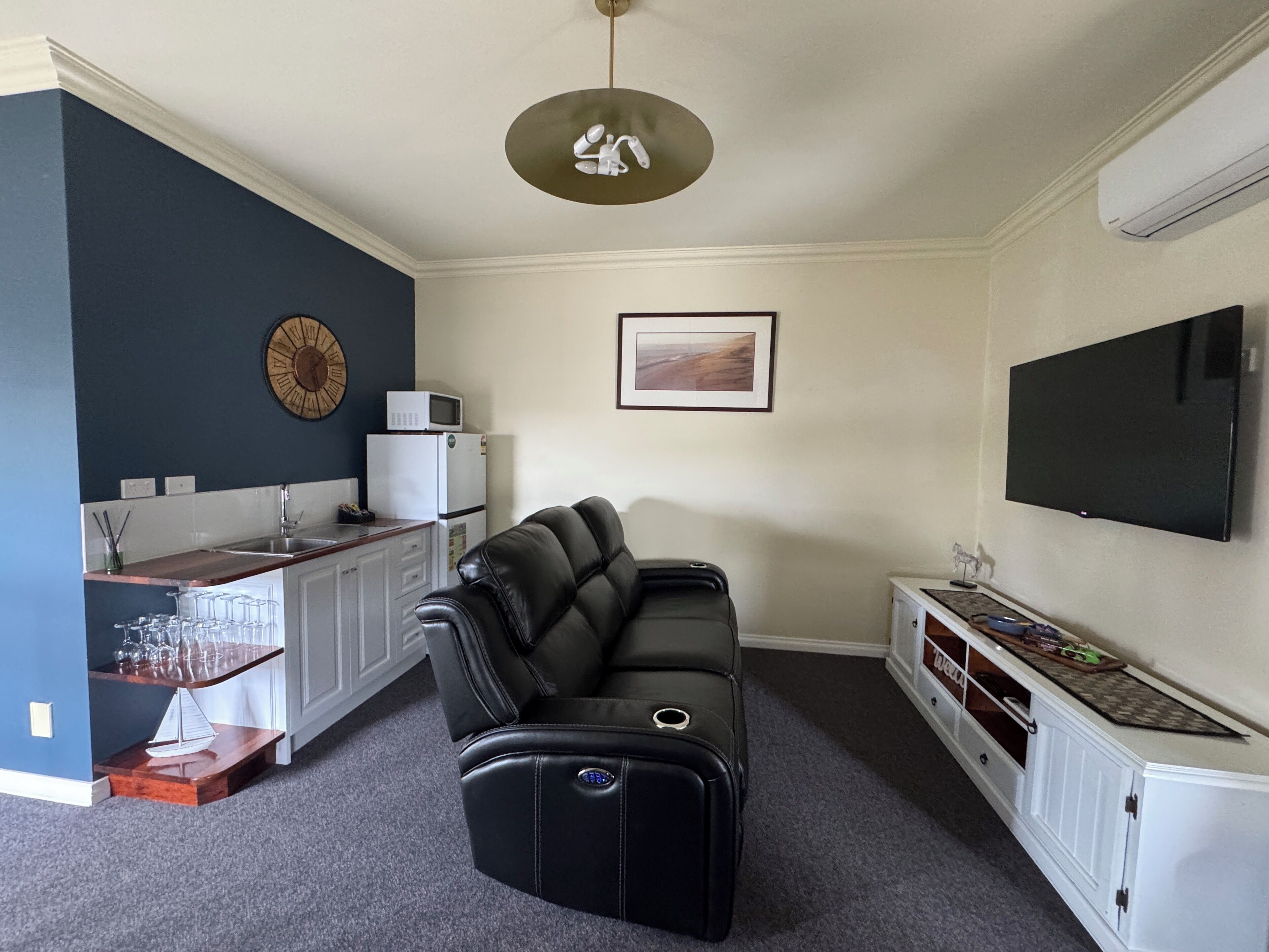 A living room with a black leather sofa, a flat-screen TV, a kitchen area with a microwave, and a ceiling fan.