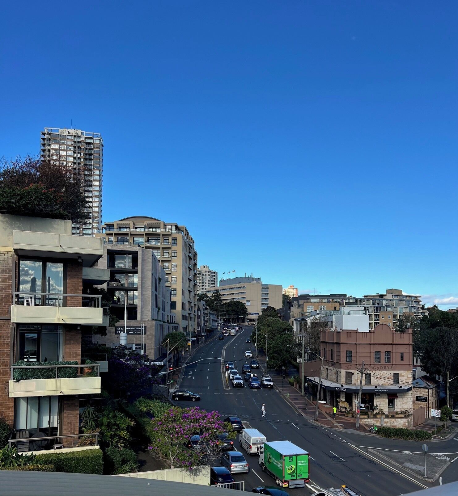 Um edifício com várias andares e varandas, uma rua com carros, e um céu azul límpido.