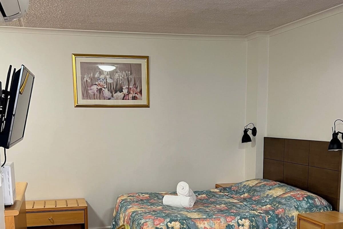 Una habitación de hotel con una cama, un escritorio, una silla, una mesita de noche, un cuadro en la pared y un aire acondicionado instalado en el techo.