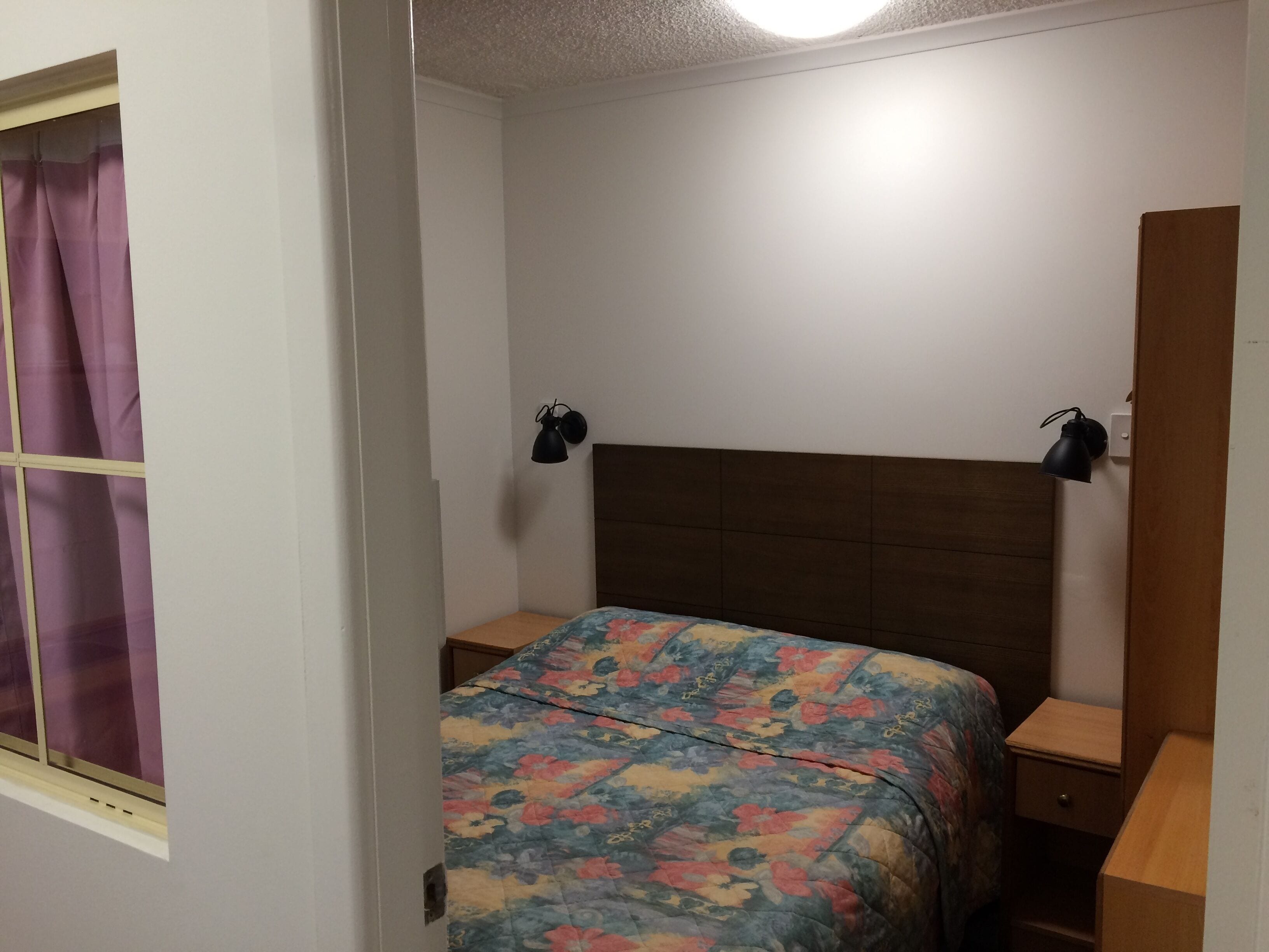 A bedroom with a bed, two bedside tables, a nightstand, a window with curtains, and two wall-mounted lamps.