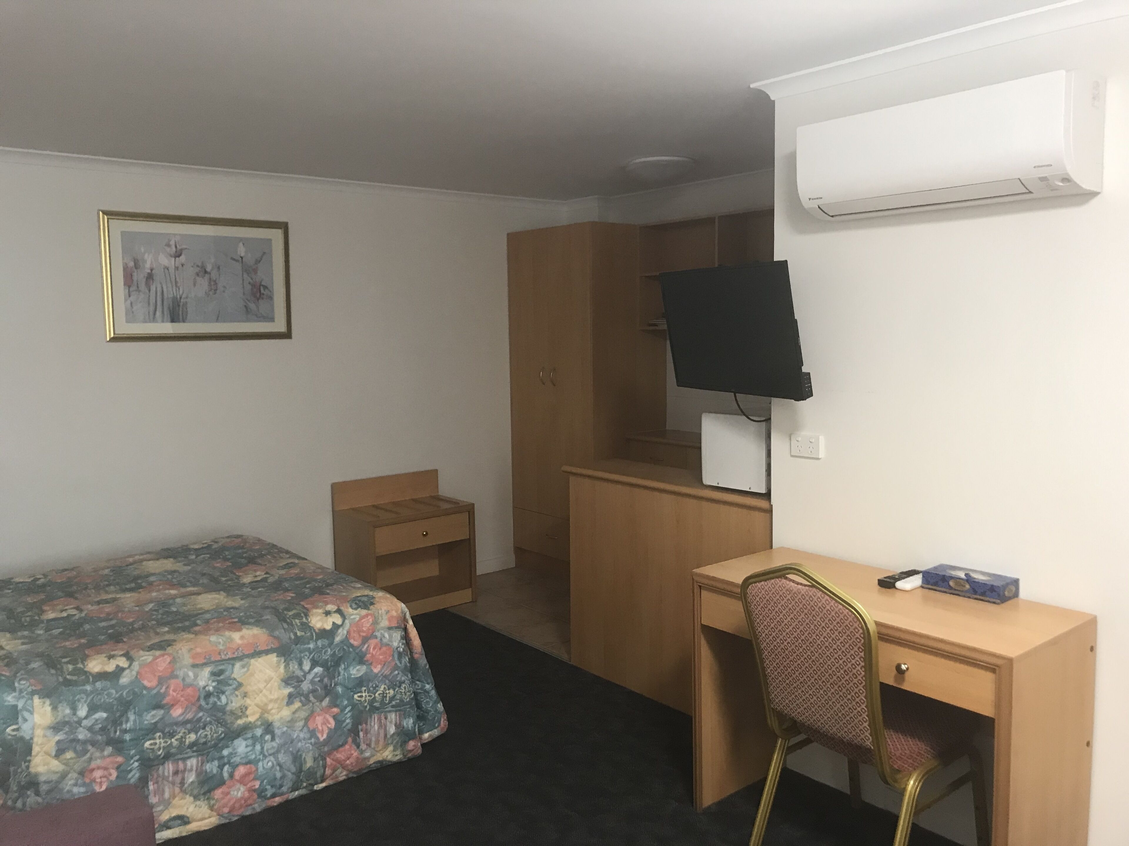Quarto de hotel com cama, escrivaninha, cadeira, televisão e guarda-roupa.