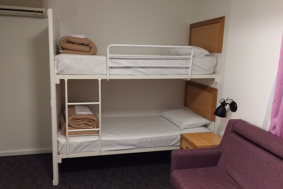 A bunk bed with a wooden headboard, a small wooden nightstand, a purple chair, and a wall-mounted lamp.