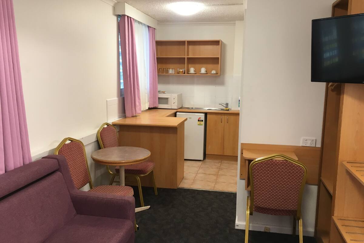 Una kitchenette compacta con microondas, pileta y placares de madera. Hay un pequeño comedor con mesa y dos sillas, y un sofá al frente.