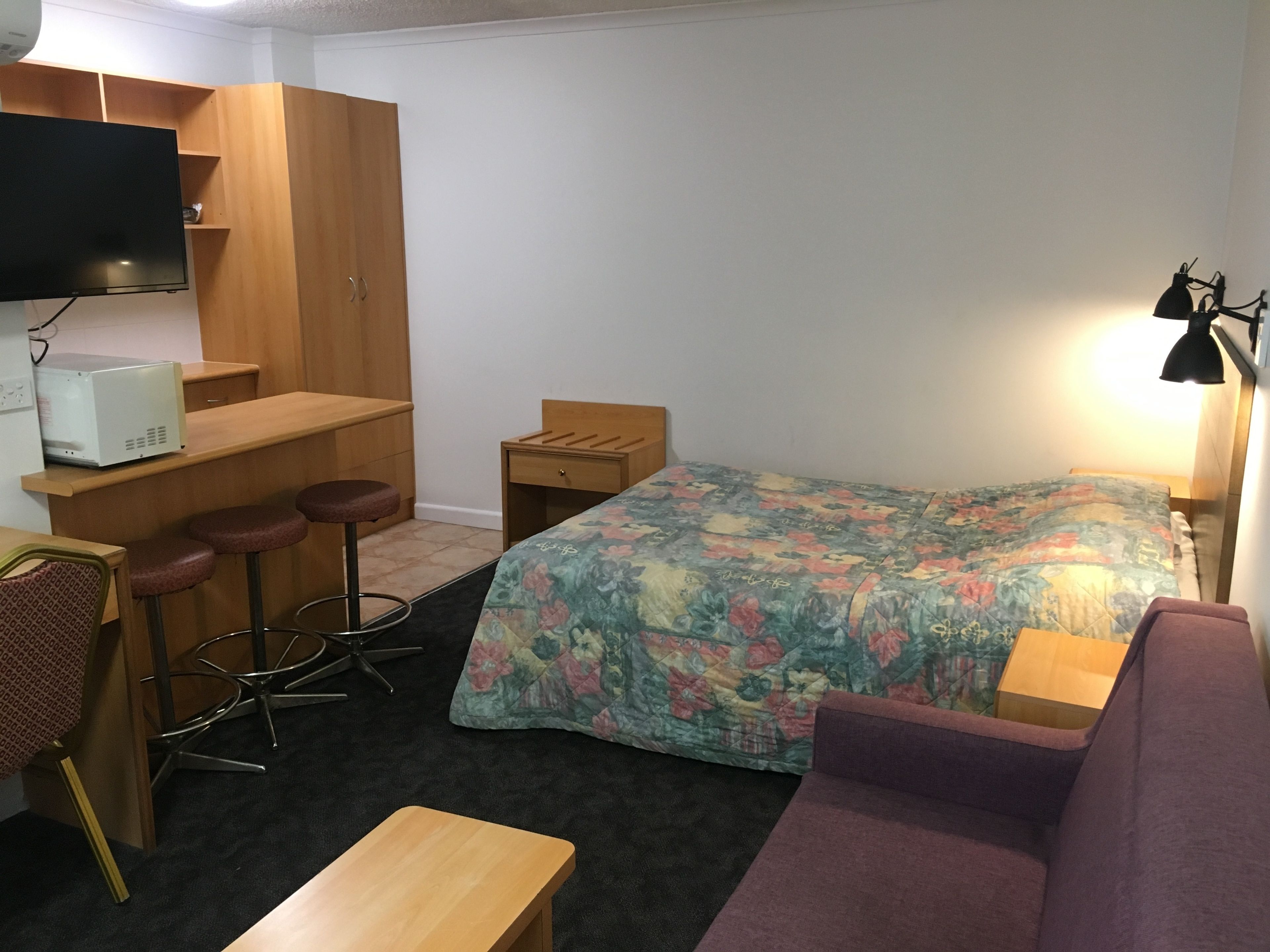 A compact hotel room with a bed, a desk with a chair, a small table, a sofa, and a TV mounted on the wall.
