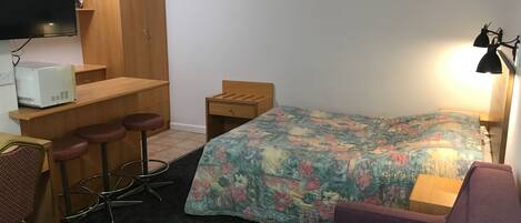 A compact hotel room with a bed, a desk with a chair, a small table, a sofa, and a TV mounted on the wall.