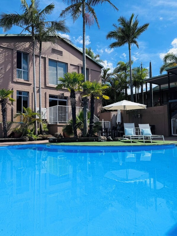 A swimming pool with lounge chairs and umbrellas, surrounded by palm trees and a building with large windows.