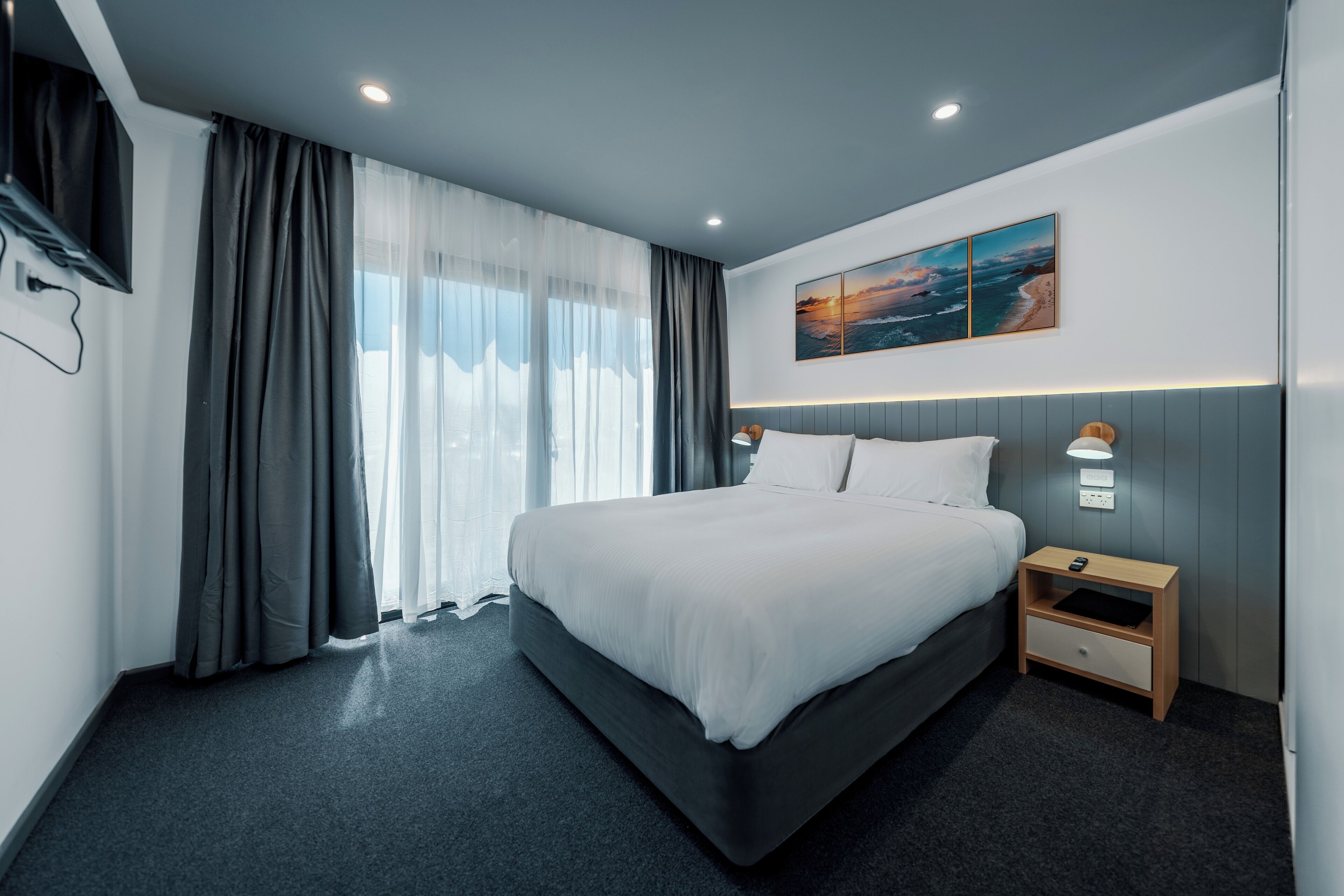 Moderni hotellihuone, jossa on suuri sänky, yöpöytä lampun kanssa ja taulu seinällä.