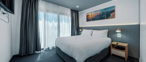 Moderni hotellihuone, jossa on suuri sänky, yöpöytä lampun kanssa ja taulu seinällä.
