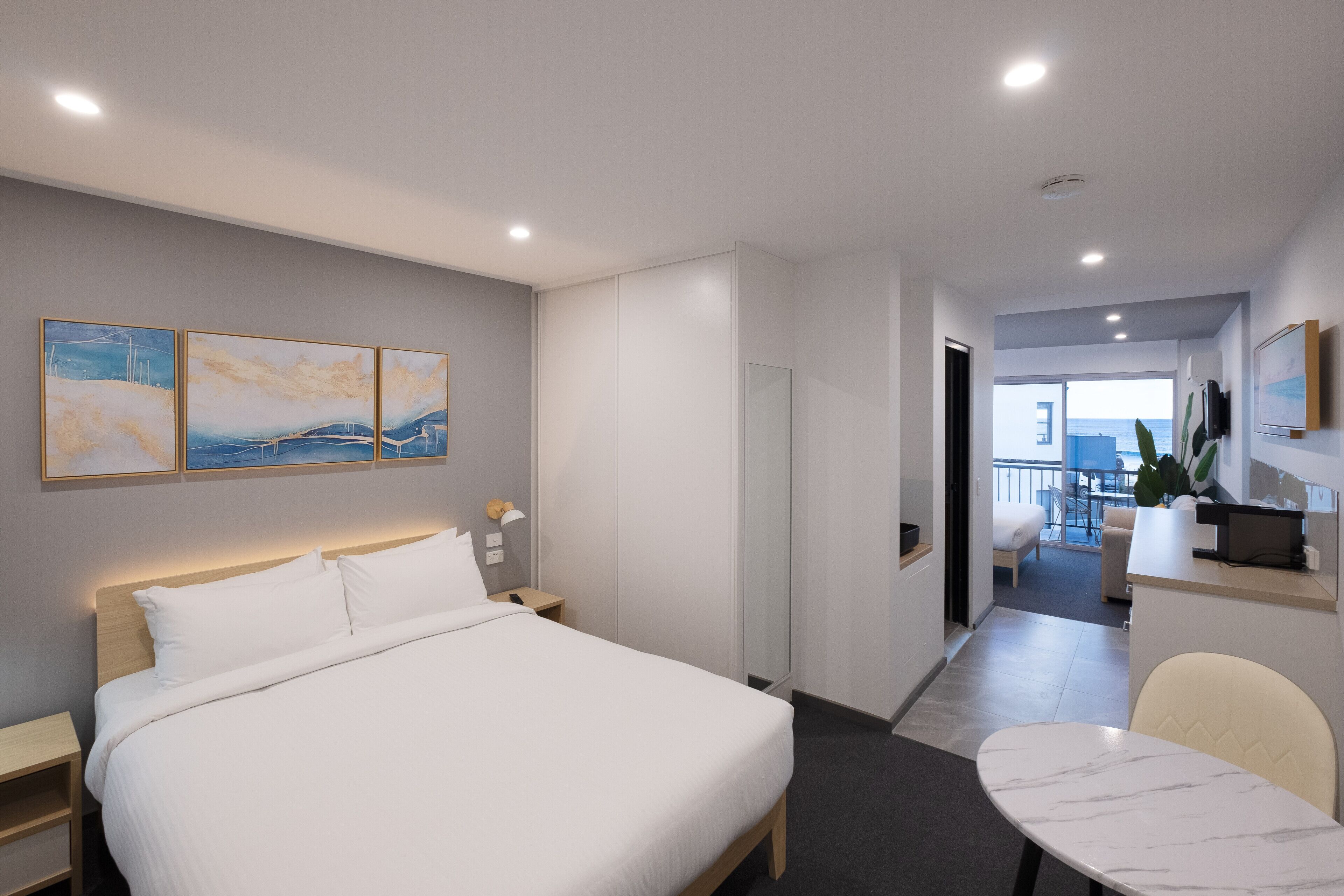 A modern hotel room with a large bed, bedside tables, a desk, and a chair. There are three paintings on the wall, a mirror, and a view of the balcony and cityscape.