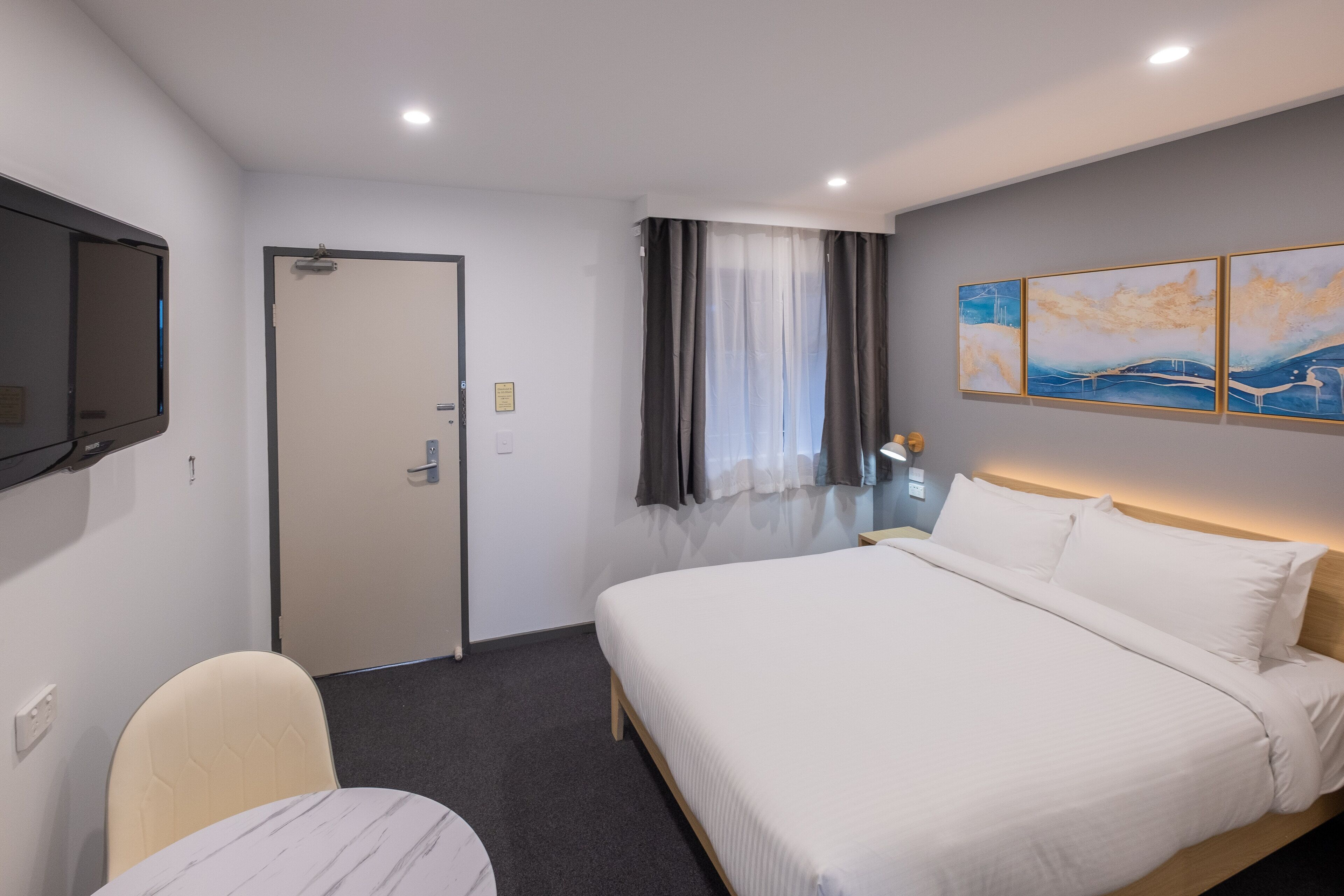 Una camera d'albergo con un letto, una sedia, una TV a schermo piatto, una porta, una finestra con tende e decorazioni alle pareti.