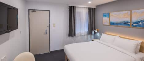 A hotel room with a bed, a chair, a flat-screen TV, a door, a window with curtains, and wall art.