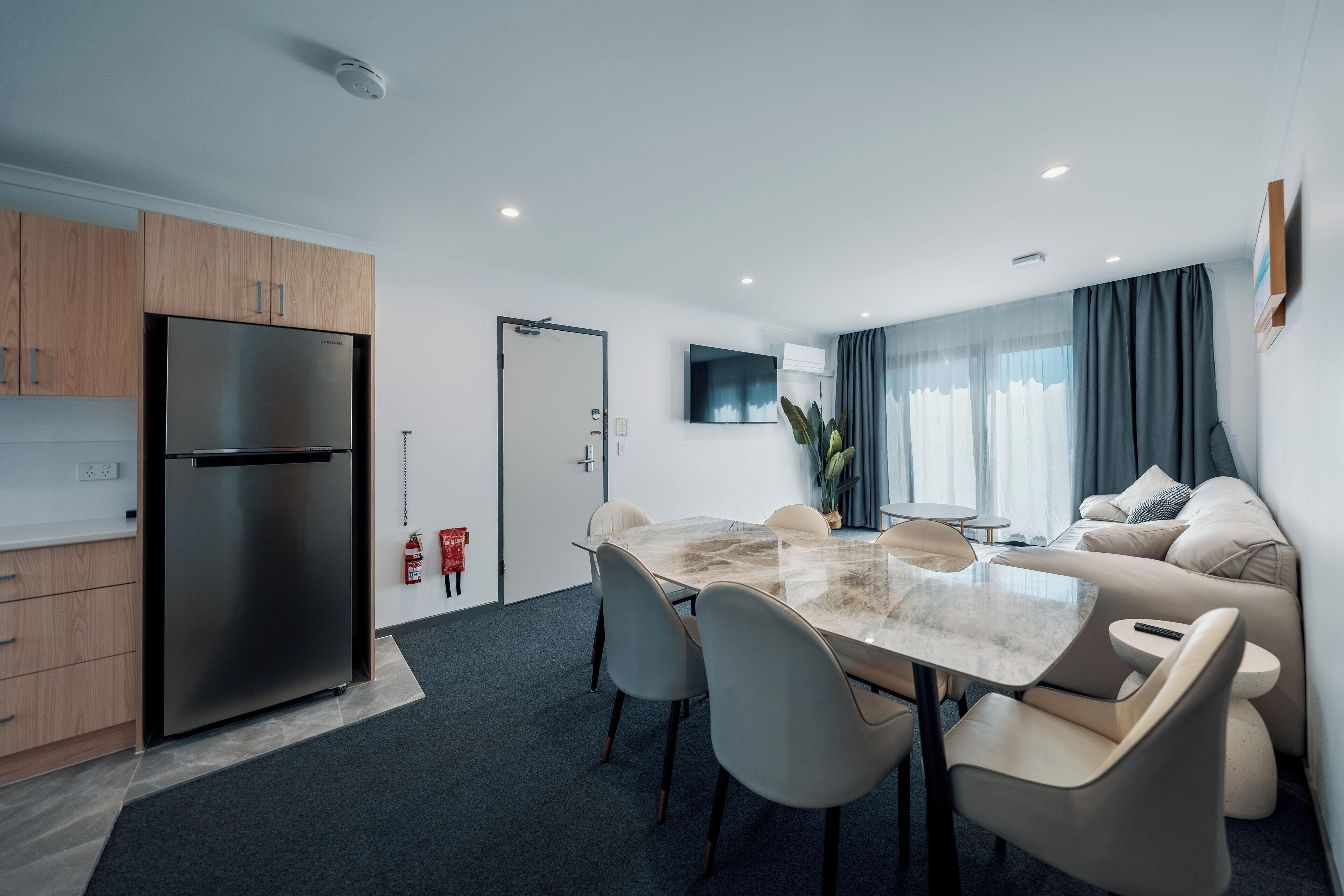 Une cuisine moderne dotée d’un réfrigérateur en acier inoxydable, un coin repas avec une table et des chaises en verre, et un salon avec un canapé et un téléviseur.