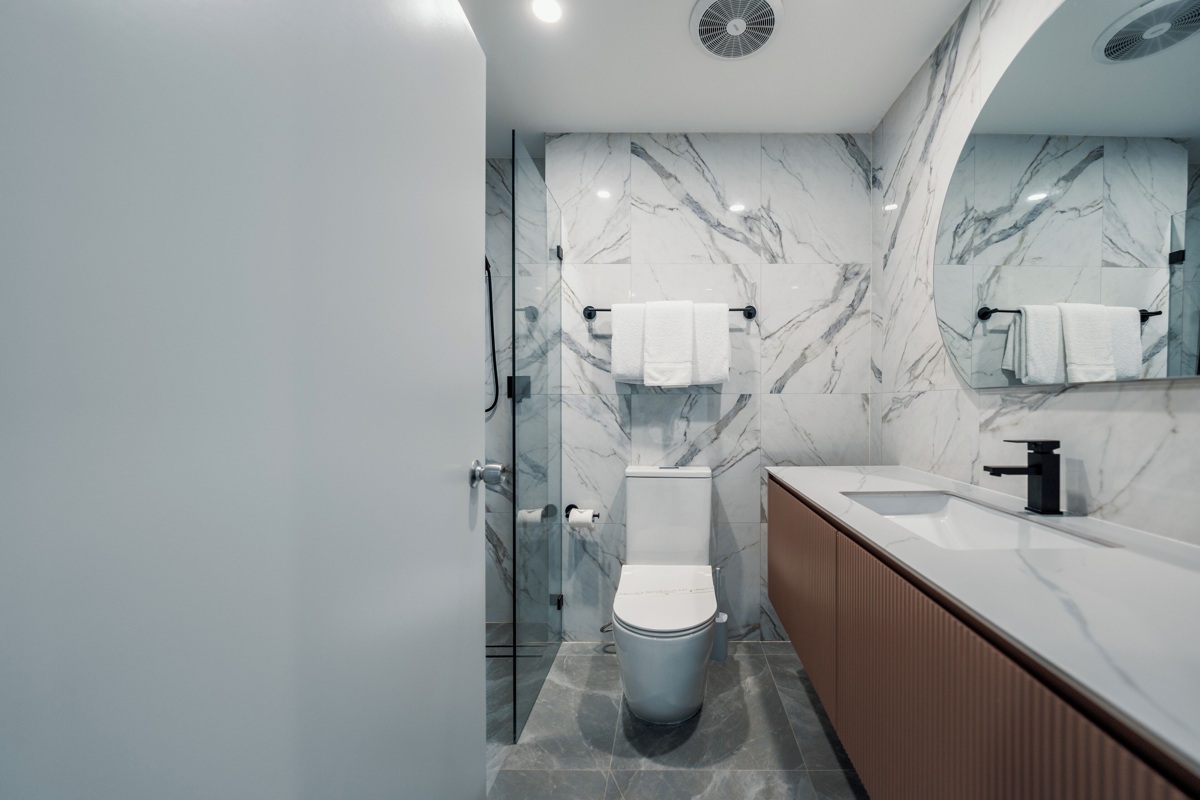 Un baño moderno con salpicadero de mármol, cabina de ducha con mampara de vidrio, inodoro blanco y tocador de madera con lavamanos.