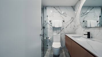 Un baño moderno con salpicadero de mármol, cabina de ducha con mampara de vidrio, inodoro blanco y tocador de madera con lavamanos.