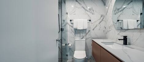 Un baño moderno con salpicadero de mármol, cabina de ducha con mampara de vidrio, inodoro blanco y tocador de madera con lavamanos.