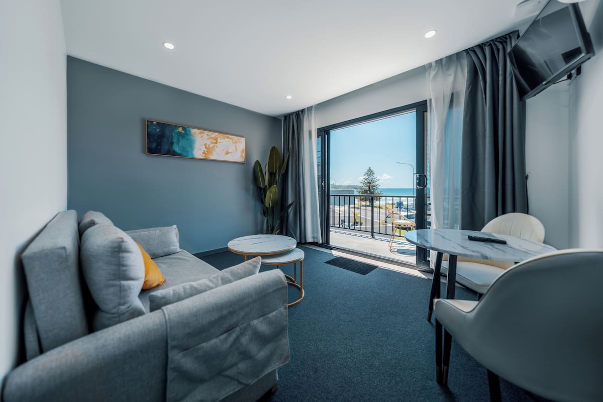 A modern hotel room with a grey sofa, a small round table, and a balcony with a view of the sea.