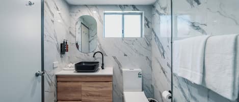 Un baño moderno con pared de mármol, tocador de madera, inodoro y cabina de ducha con mampara de vidrio.