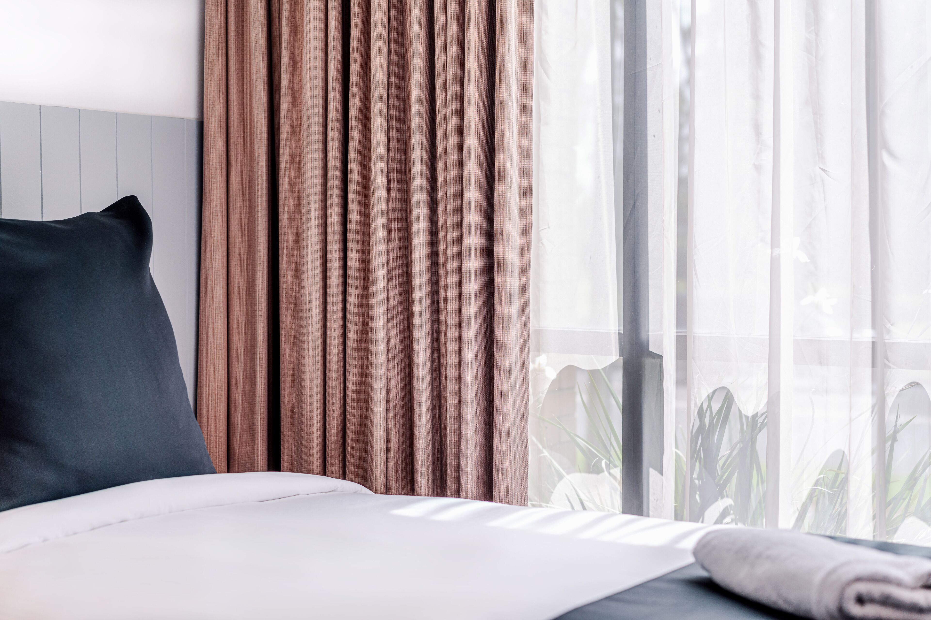 Una cama bien hecha con almohada oscura y sábanas blancas, una ventana con cortinas translúcidas y vistas al verdor exterior.