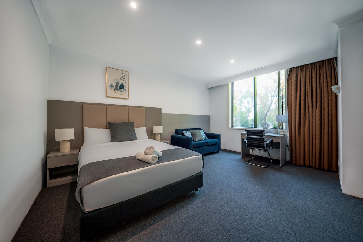 Una habitación de hotel moderna con una cama grande, un escritorio, un sofá azul y una ventana con cortinas.
