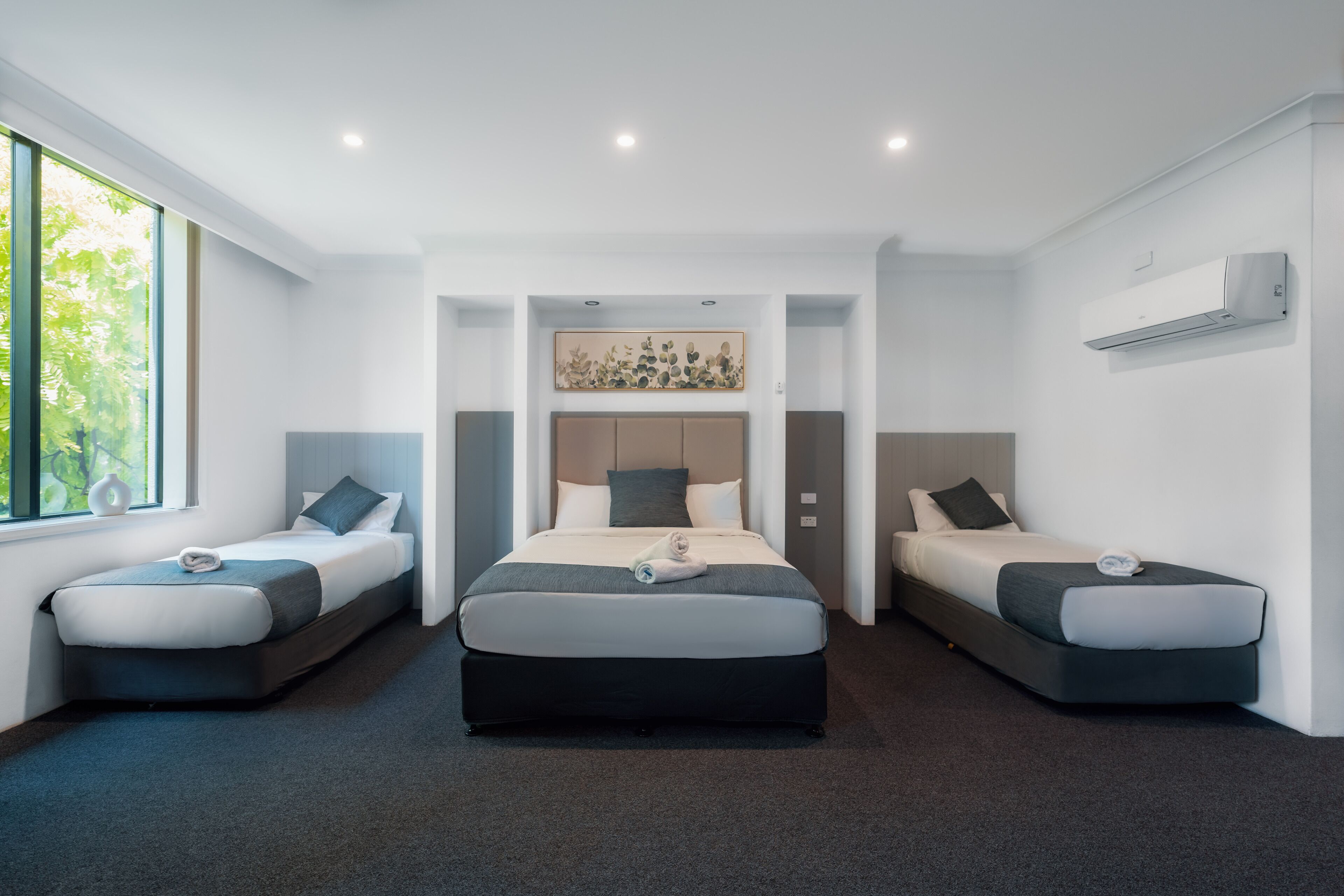 Una habitación de hotel moderna con una cama central flanqueada por dos camas más pequeñas, un ventanal grande y un equipo de aire acondicionado en la pared.