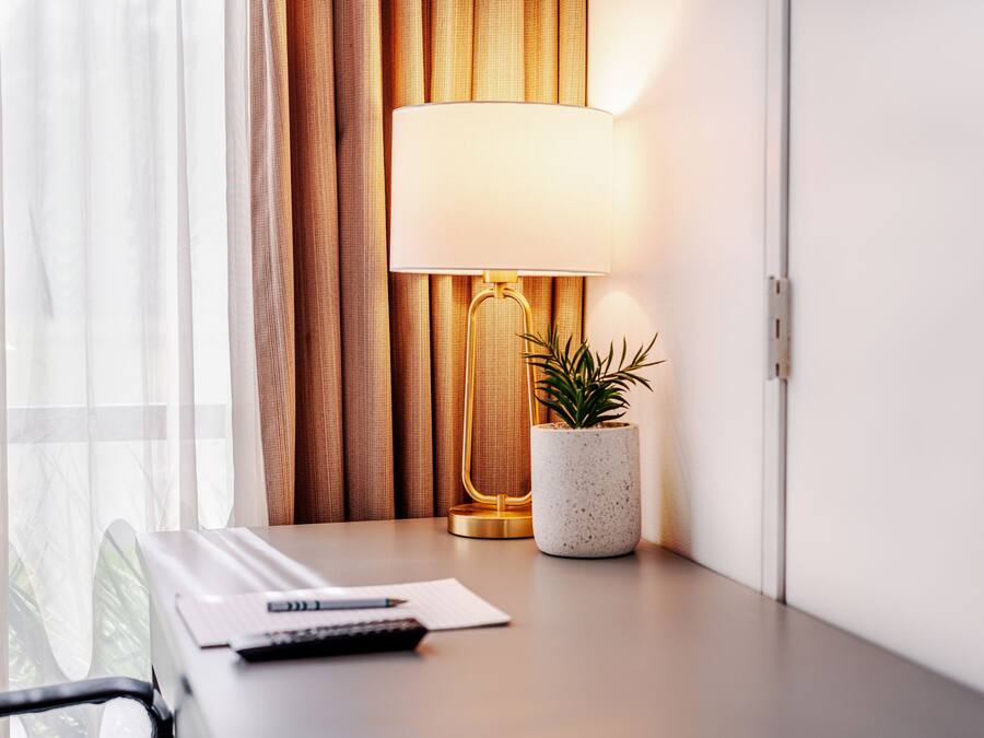 Hotellihuone, jossa on työpöytä, lamppu, ruukkukasvi ja verhot.
