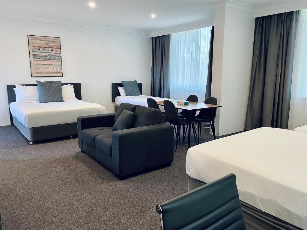Een hotelkamer met twee bedden, een bank, een eettafel met stoelen en een groot raam met gordijnen.